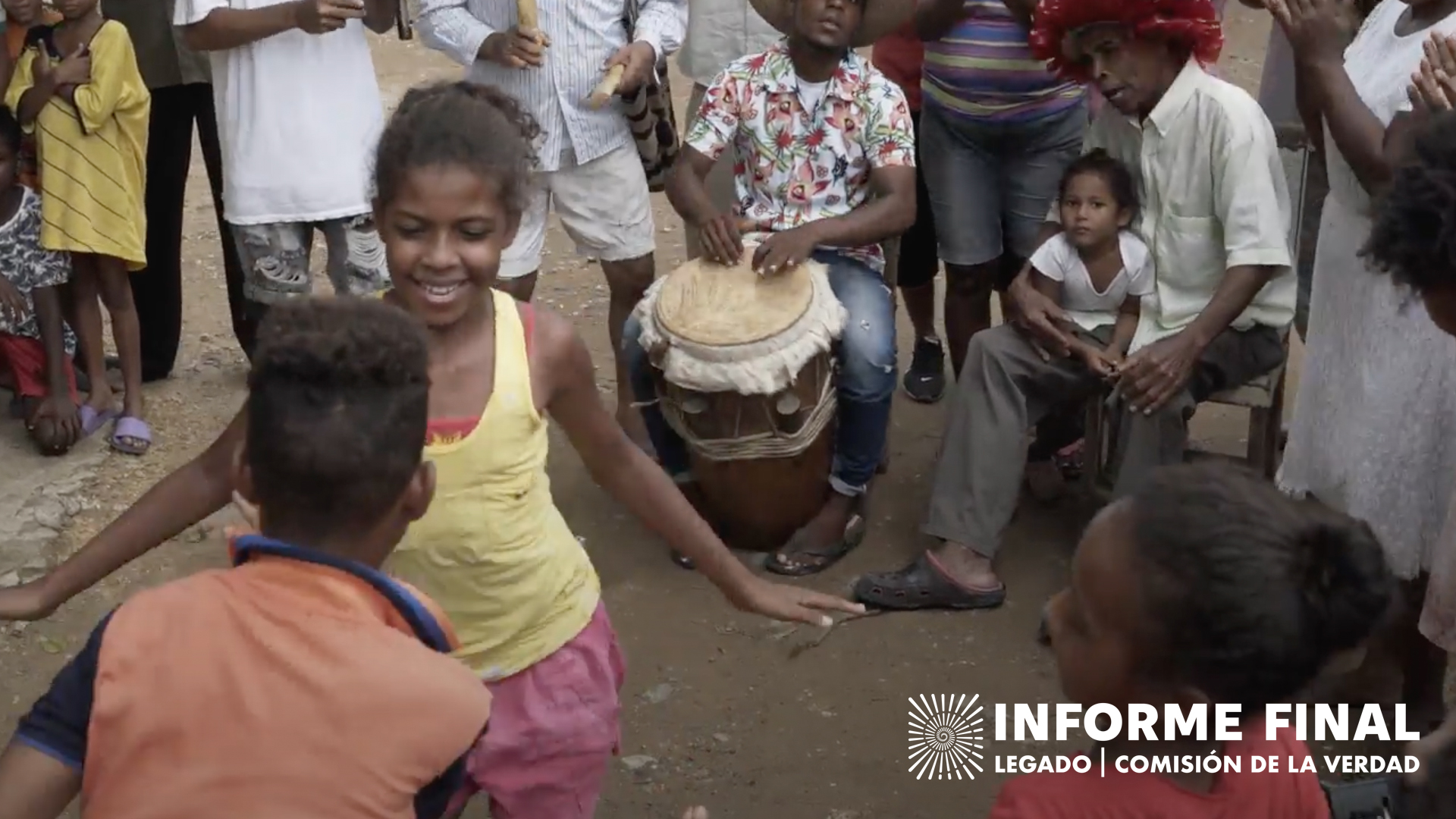 Niña afro bailando y sonriendo con un niño dentro de un círculo de personas, un hombre toca tambor