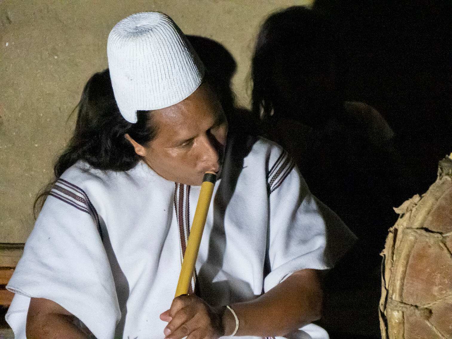 En la noche, un hombre arhuaco sentado y vestido con traje tradicional, toca un instrumento de aire.