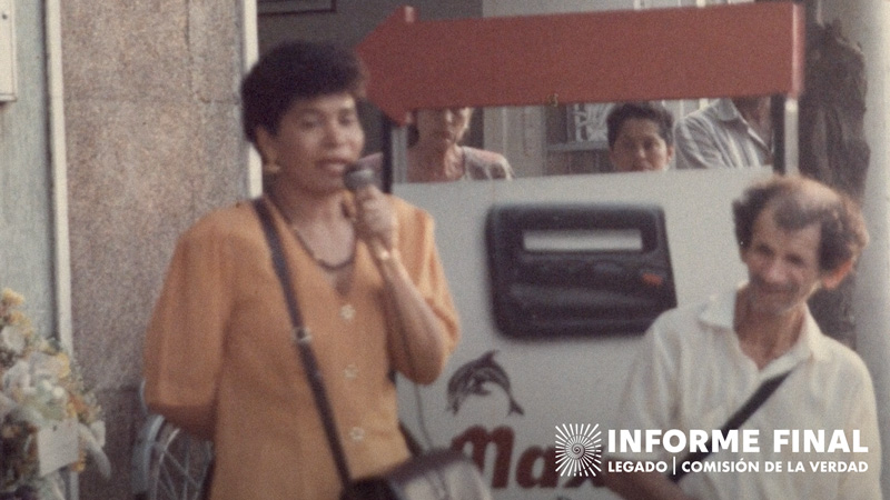 Captura fotográfica antigua, mujer con bolso habla por micrófono que sostiene, le rodean personas atentas 