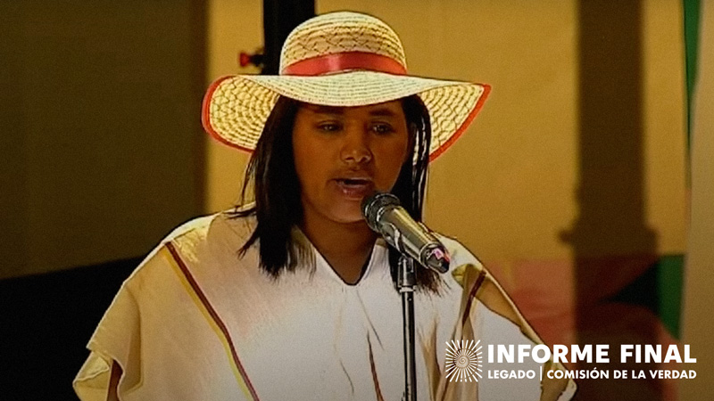 Claudia Machuca hablando frente a micrófono, porta sombrero tejido y poncho blanco