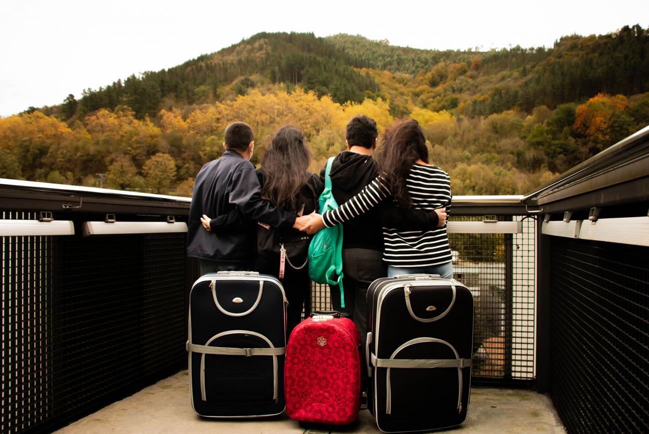 frente a tres maletas de viaje, cuatro personas de espaldas se abrazan y miran una montaña