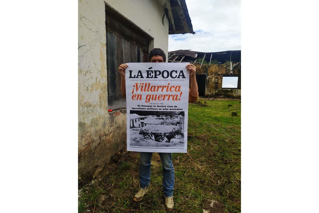 Un hombre oculta su rostro con un afiche. texto: “La época. Villarrica en Guerra. Fondo con casa abandonada y cielo nublado