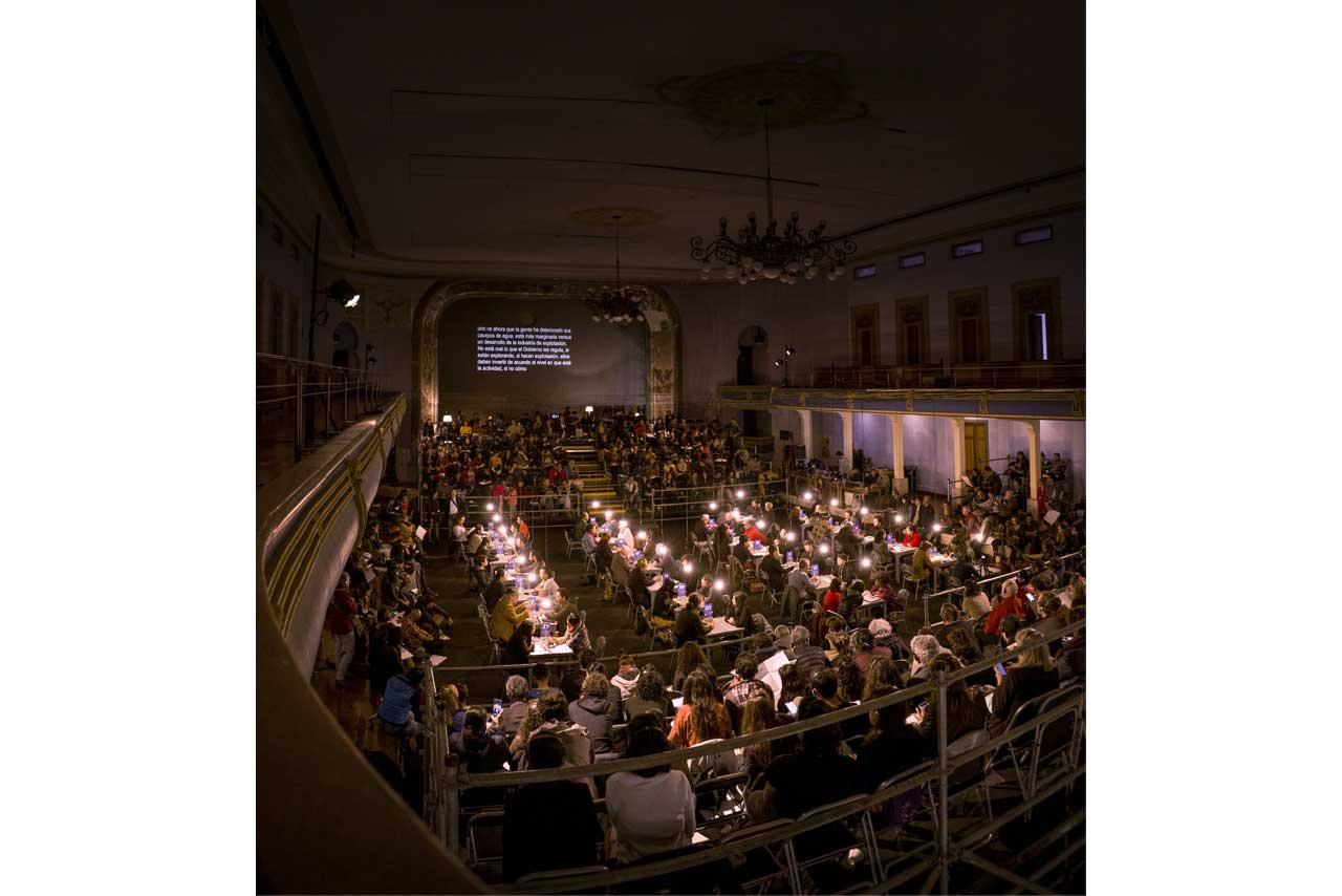 Plano general del evento. Teatro antiguo, cientos de asistentes, mesas con luz baja en el centro, personas conversando