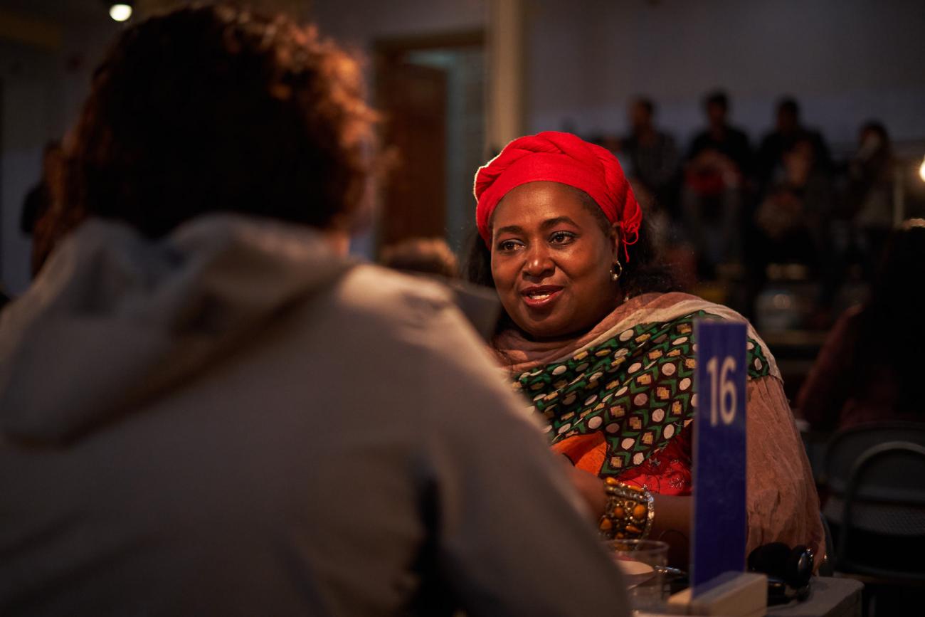 Mujer afro con ropa colorida conversa con otra persona en mesa número 16, luz cálida
