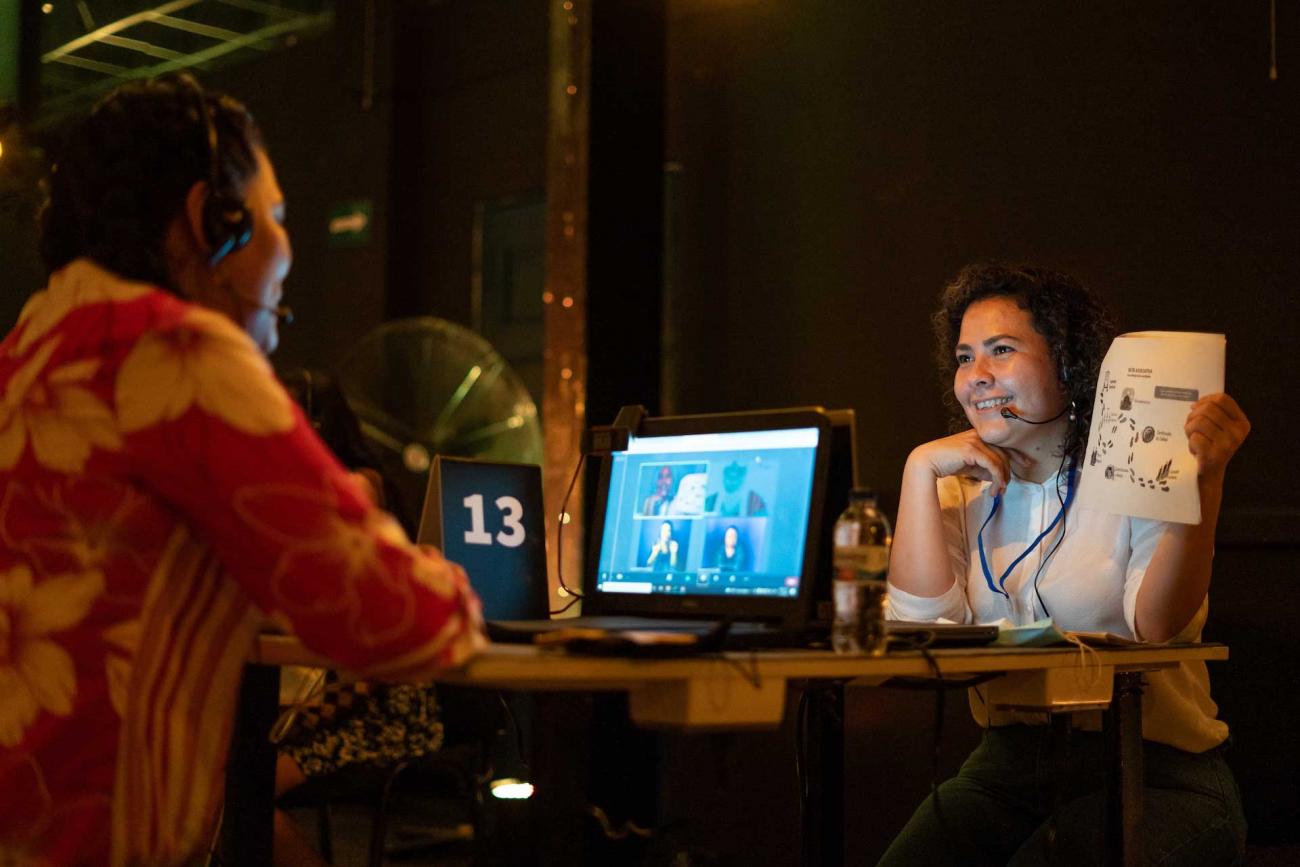 Mesa trece, dos mujeres comentan una gráfica impresa, en la pantalla del computador se ven más personas en conferencia