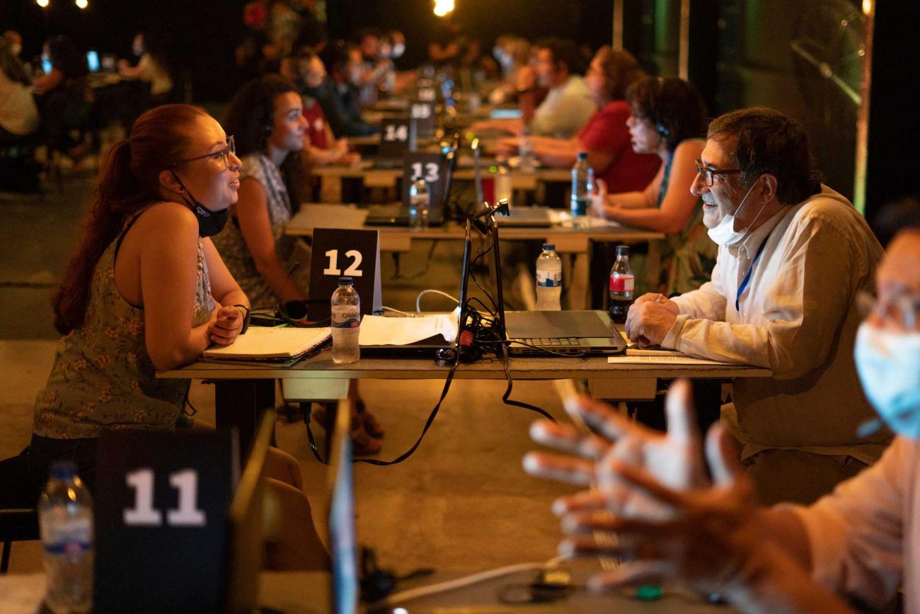 Hombre y mujer conversan frente a frente en una mesa. En otras mesas, parejas de personas dialogan