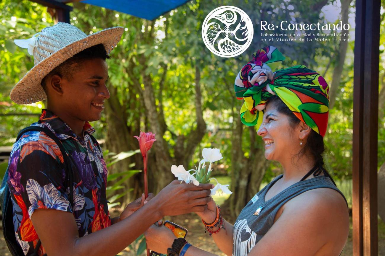 Joven con sombrero y mujer con turbante colorido intercambian flores mientras sonríen en un paisaje soleado y con árboles