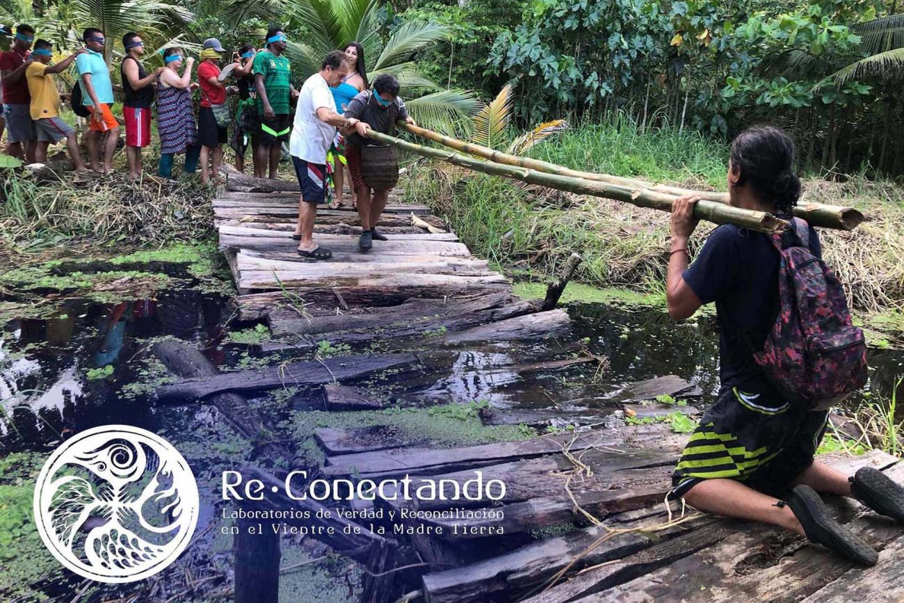 Un grupo con ojos vendados pasa por un puente de madera colapsado sobre un riachuelo. dos hombres sostienen guaduas a modo de barandas