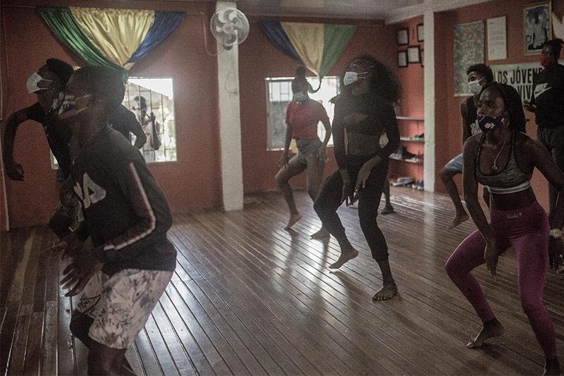  Mujeres y hombres afro con tapabocas bailan en un salón de baile, un hombre los mira desde una ventana