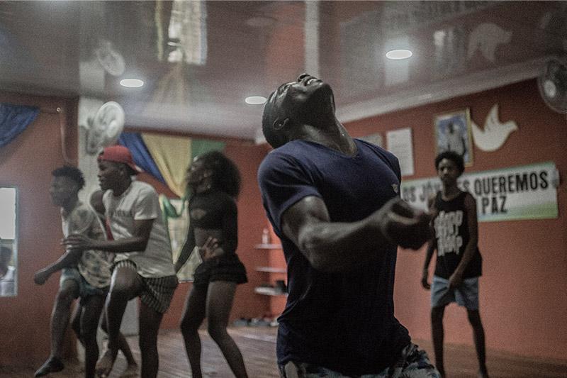 Un hombre afro baila y mira al techo, detrás de él jóvenes bailan con ropa deportiva