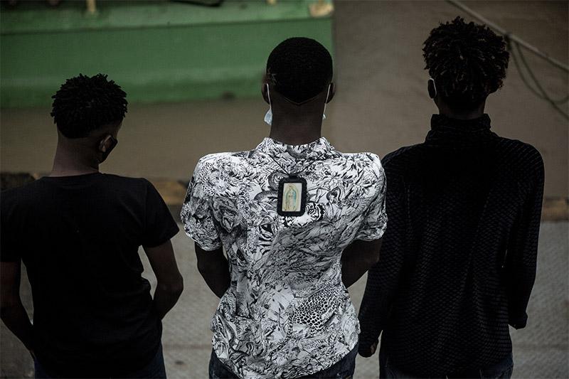 Tres hombres afro de espalda, dos visten de negro y el del medio con camisa blanca estampada, tiene una imagen de la virgen en la espalda