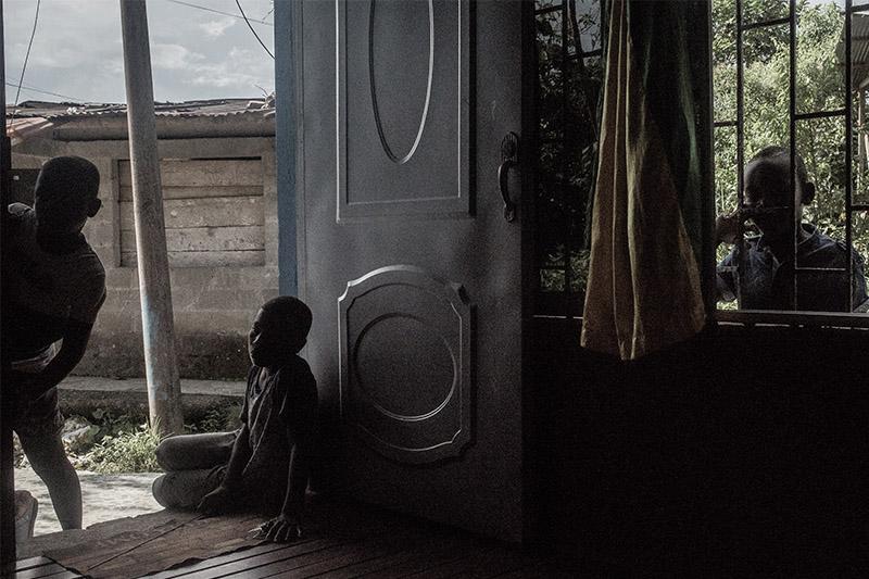Dos niños afro están asomados desde afuera por una puerta de metal de una casa con piso de madera, otro niño mira hacia adentro por una ventana