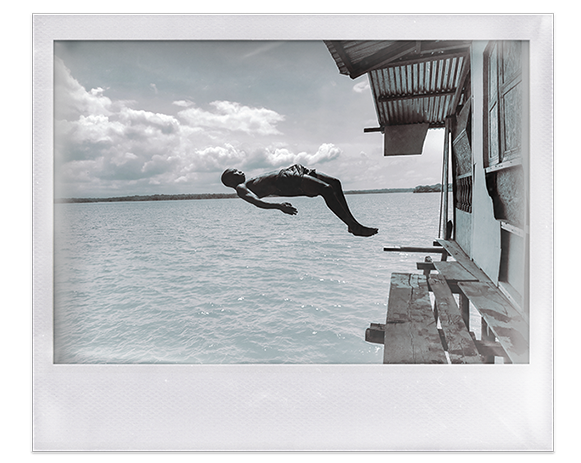 Instantánea. Hombre afro en pantaloneta salta de espaldas, desde un palafito, al mar