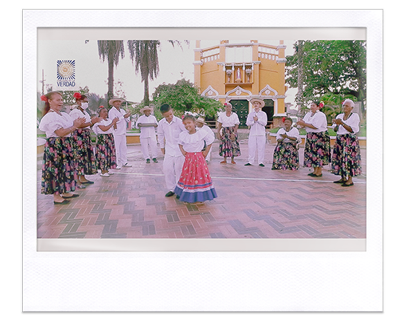Instantánea. Niña y niño con vestidos folclóricos bailando en una plaza, rodeados por personas igualmente vestidas 