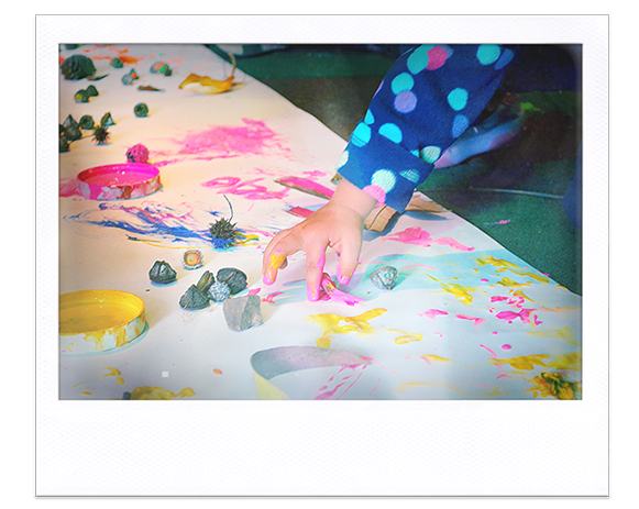 Instantánea. Mesa con semillas y papel, niño juega con pinturas haciendo manchas con sus dedos