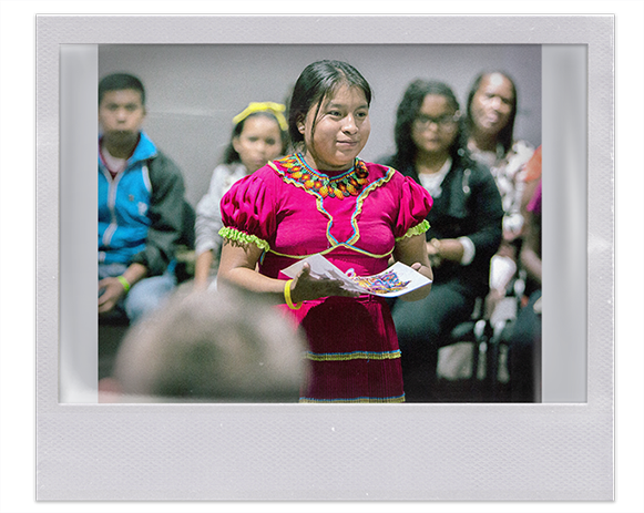 Instantánea. Joven mujer indígena con vestido de colores vivos y collar, en sus manos porta una ofrenda de pulseras