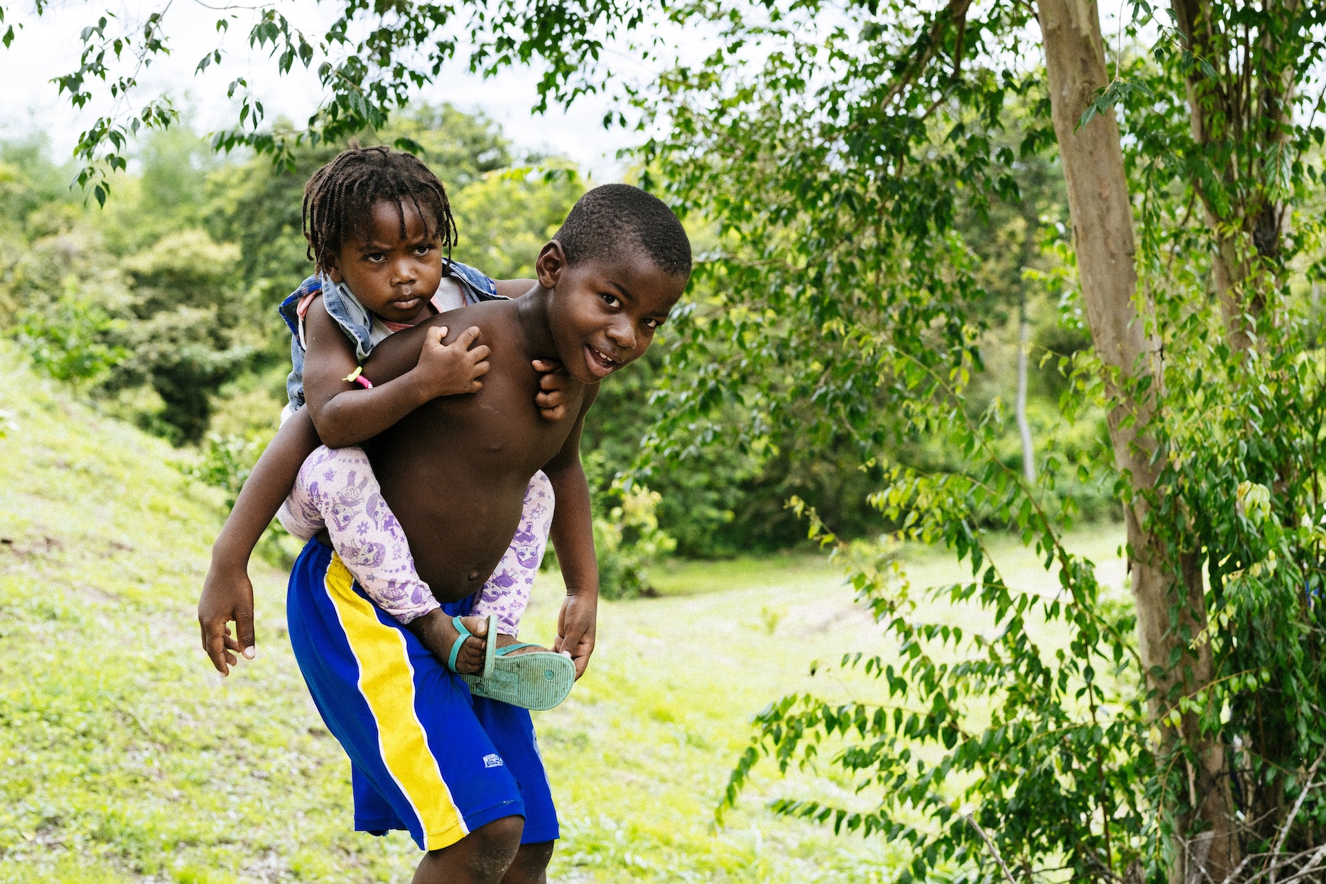 Niño en pantaloneta y sin camiseta carga a una niña de trenzas sobre su espalda, al fondo árboles