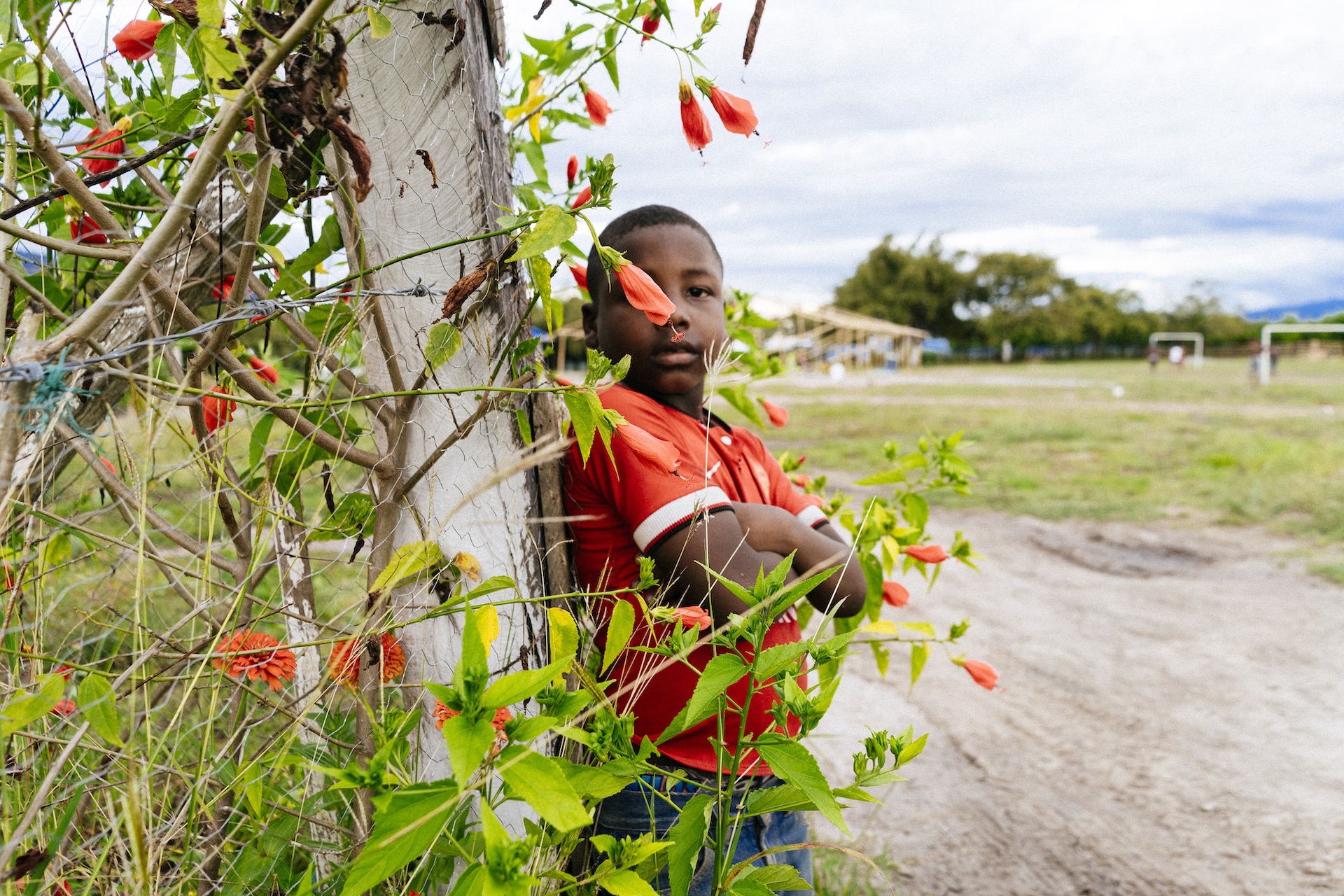 Niño de brazos cruzados, arbusto con flores, cancha de fútbol y casa de madera en construcción