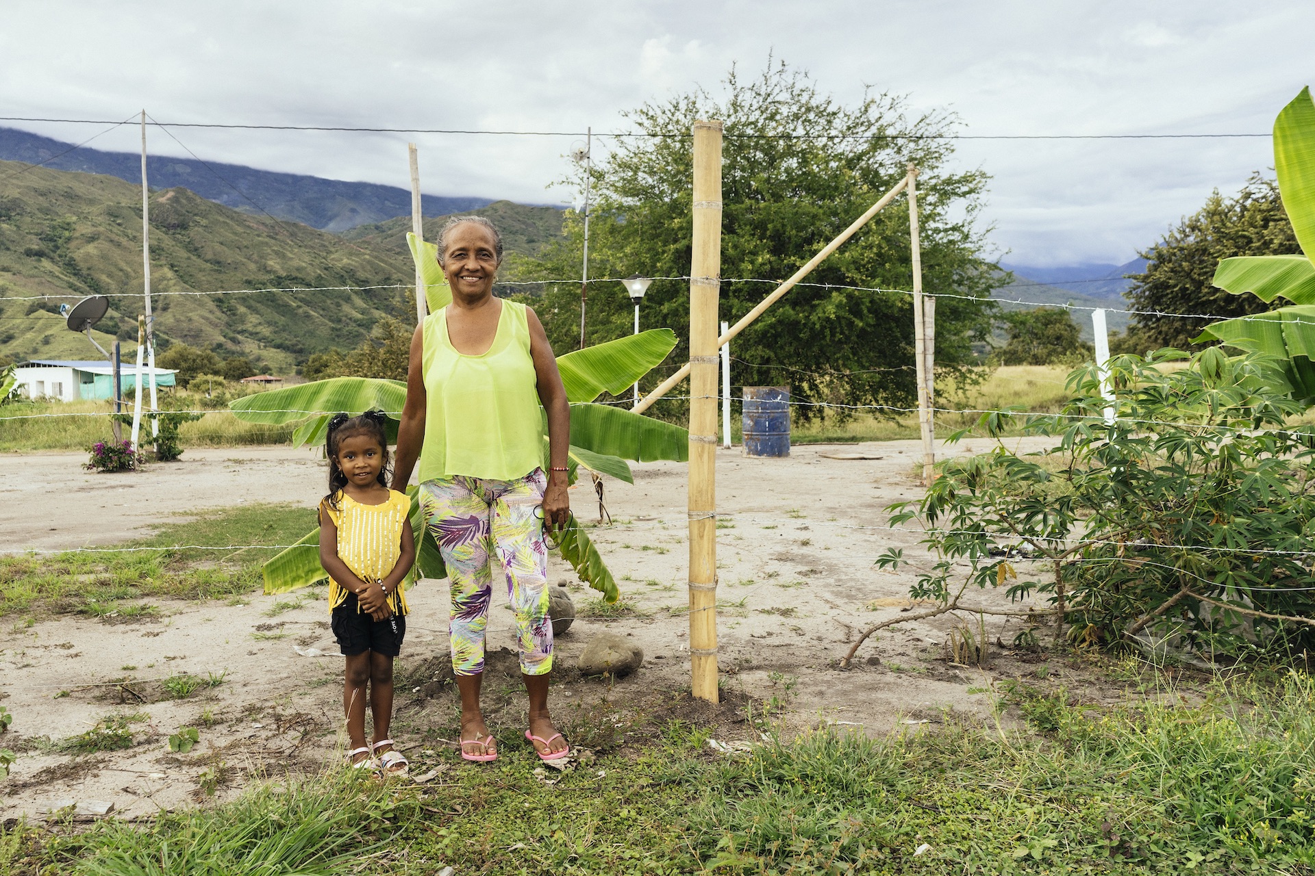 Mujer con una niña sonriendo, detrás una cerca de alambre de púas, planta de plátano y montañas