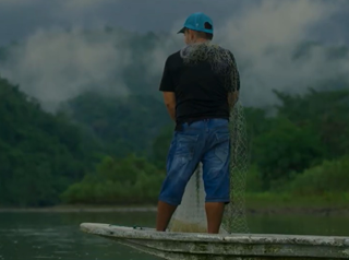 Hombre montado en una lancha, sosteniendo una red. Está de espaldas mirando el Río Sogamoso y de fondo se ven montañas